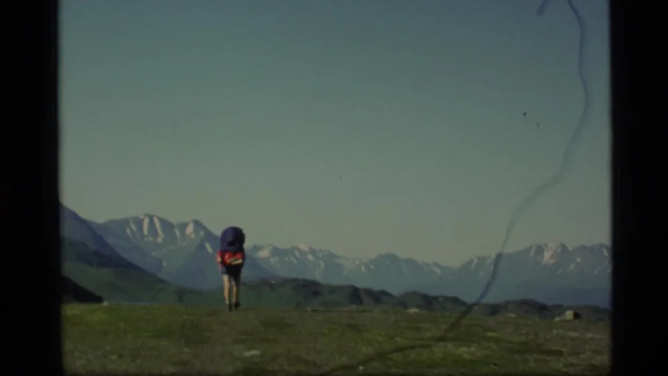 A backpacker walks away across open alpine grass in Alaska, with distant snowy peaks and visible 8mm film wear near the frame edge.