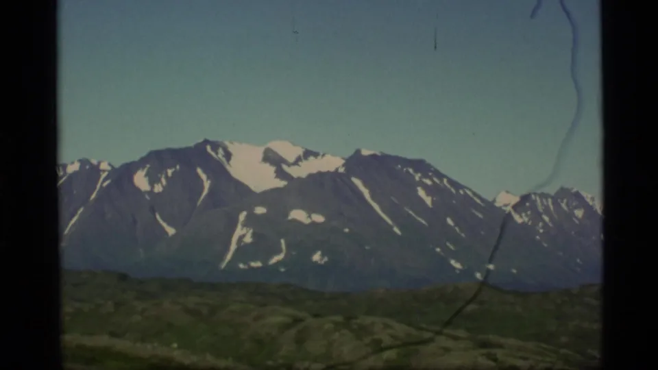 Snow-capped Alaska mountains fill the frame above green ridges, with 8mm film borders and a visible scratch from the scan.