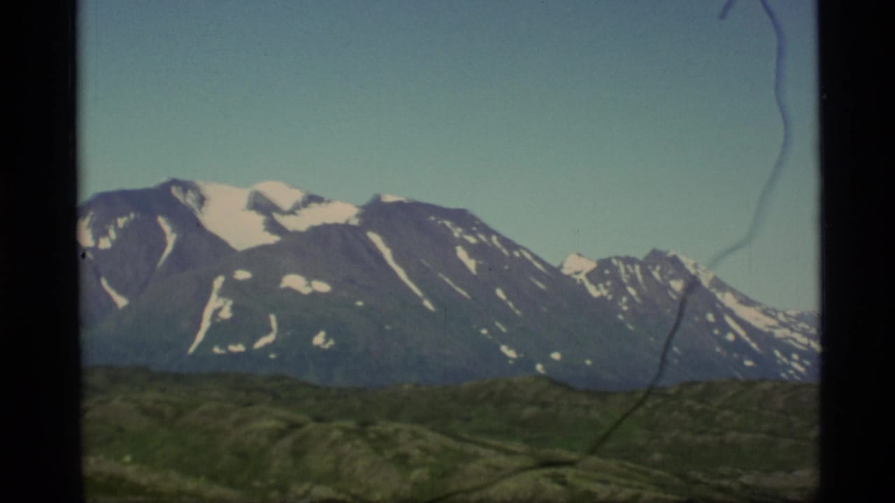 1977 Alaska 8mm frame showing green hills and distant snow-capped mountains beneath a pale blue sky.