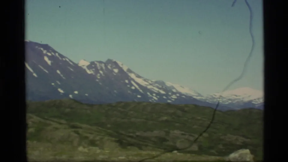 1977 Alaska home-movie frame with a broad mountain ridge, snow patches, green foreground hills, and visible film border.
