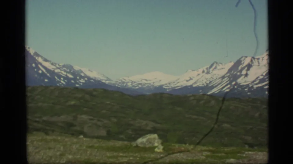 1977 Alaska 8mm frame showing distant snow-covered peaks, green rolling terrain, and a foreground rock.