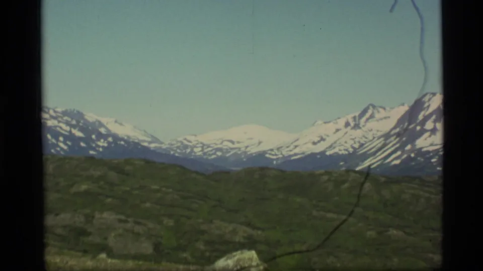 1977 Alaska home-movie frame with snow-capped mountains across the horizon and green hills in the foreground.