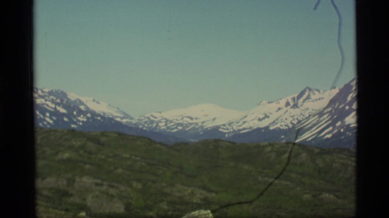 1977 Alaska 8mm frame showing distant snow-capped mountains across green open terrain under clear sky.