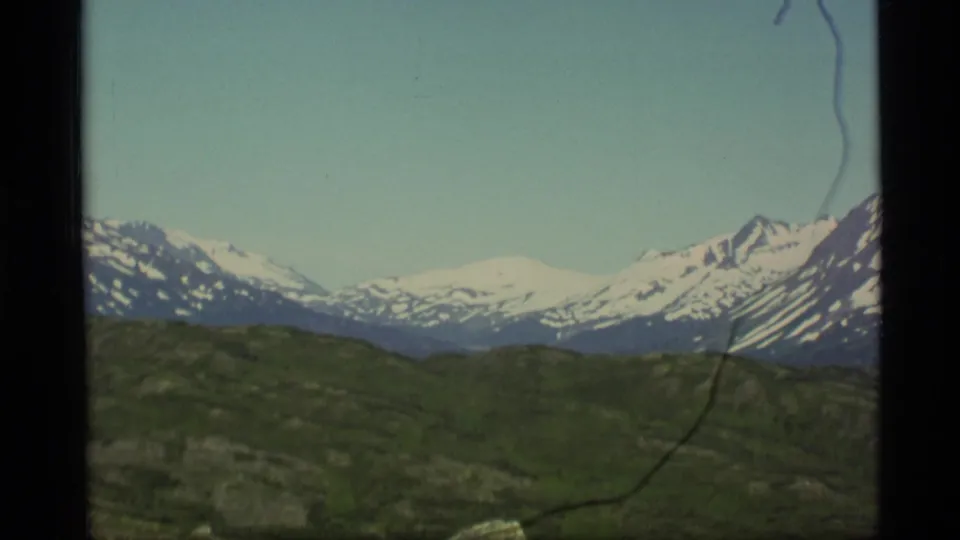 1977 Alaska 8mm frame showing distant snow-capped mountains across green open terrain under clear sky.