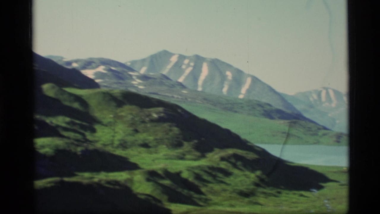 Green hillside and blue lake below snow-streaked mountains.