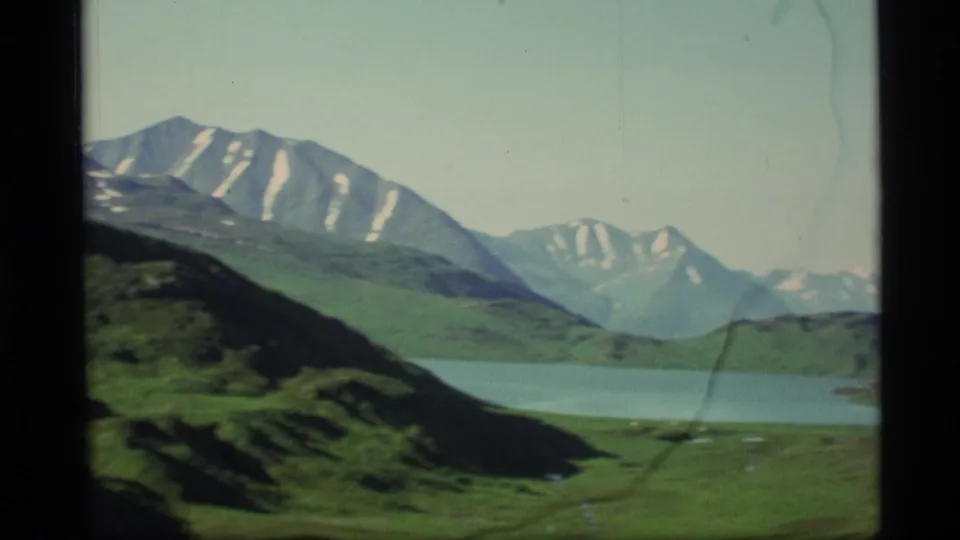 1977 Alaska 8mm frame with a blue lake, grassy valley floor, dark hillside, and distant alpine peaks.