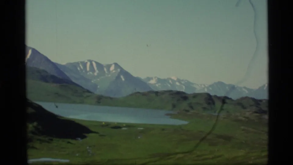 1977 Alaska 8mm landscape showing open grassland, a blue lake, and snow-marked mountains across the horizon.