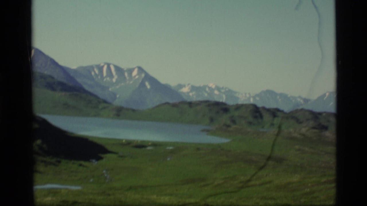 1977 Alaska 8mm frame showing a calm lake, green slopes, distant snow-streaked mountains, pale sky, and visible film wear.