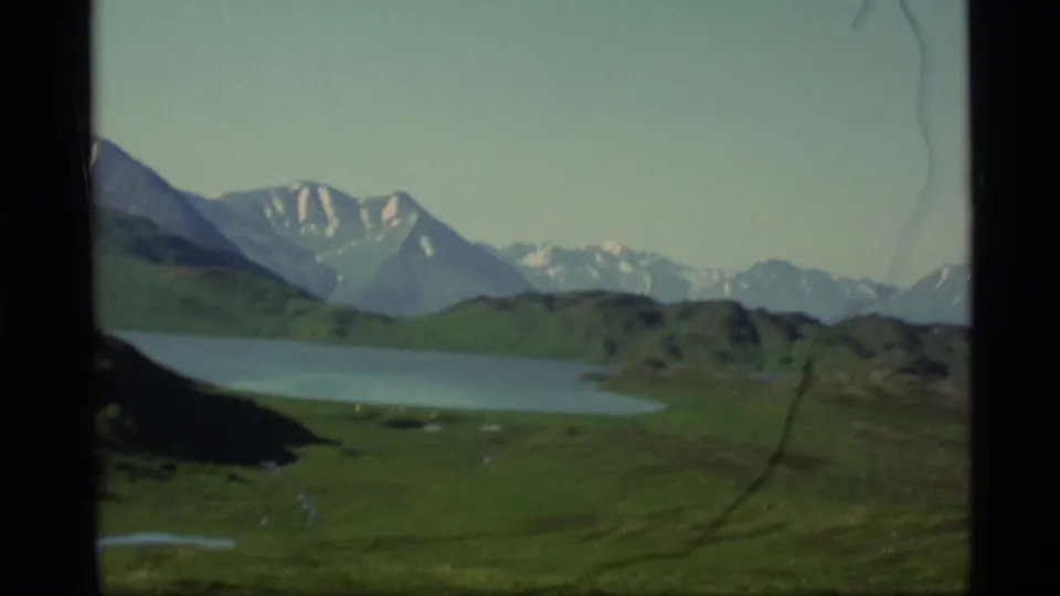1977 Alaska 8mm frame showing a calm lake, green slopes, distant snow-streaked mountains, pale sky, and visible film wear.