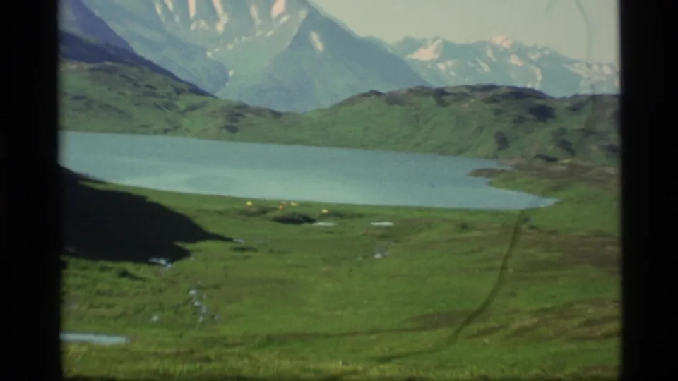 1977 Alaska home-movie frame with a blue-green lake, grassy shore, distant mountains, and small yellow shapes near the waterline.