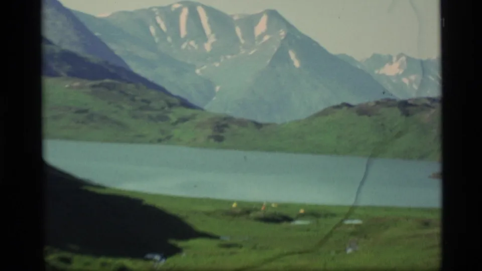 1977 Alaska 8mm frame showing the lake closer in view, green hills, high snow-streaked peaks, and scratches across the film image.