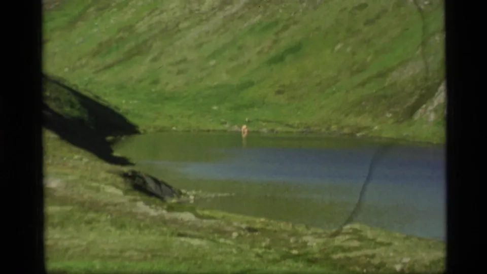 1977 Alaska 8mm frame showing a green hillside, lake edge, dark slope shadow, and a tiny distant human figure or object near the shoreline.