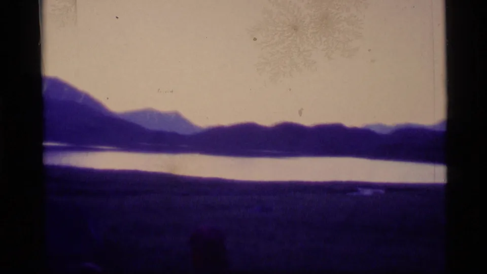 1977 Alaska home-movie frame of a quiet lake at dusk, with dark ridges and a purple-tinted foreground.