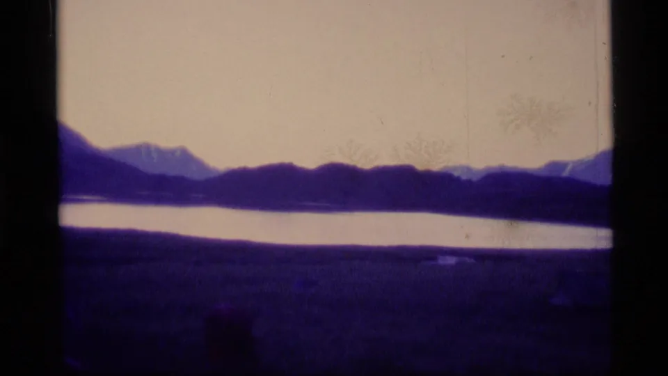 1977 Alaska 8mm frame showing a dark mountain ridge beyond calm water, with purple color shift and visible film gate borders.