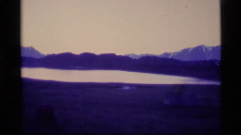 1977 Alaska home-movie frame with distant snow-capped peaks, dark ridge line, calm water, and strong purple film coloration.