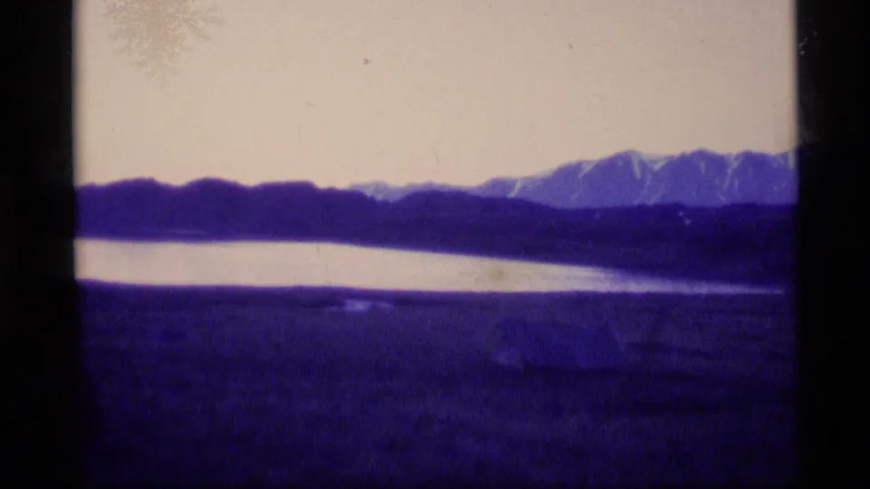 1977 Alaska 8mm frame showing bright water below a dark ridge, with snow-covered peaks entering at the right side.