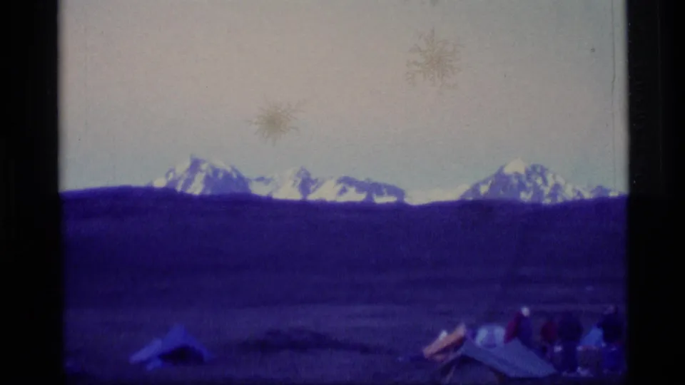 1977 Alaska 8mm frame with distant mountains, a wide field, and small campsite tents entering the lower foreground.