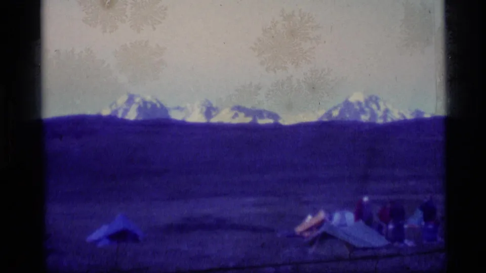 1977 Alaska 8mm frame showing snowy peaks, a broad field, tents, and small unidentified people at a campsite.
