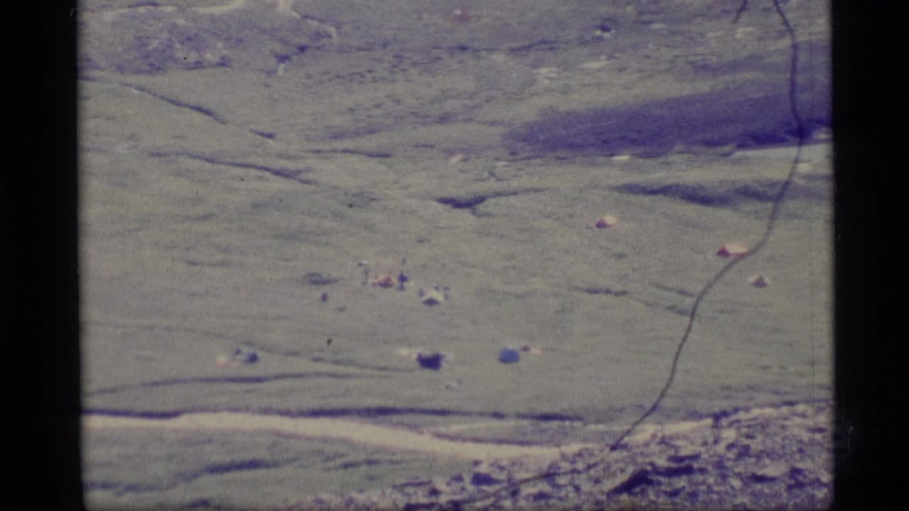 1977 Alaska 8mm aerial view of open green terrain with small tent-like forms, scattered dark marks, and visible film border.