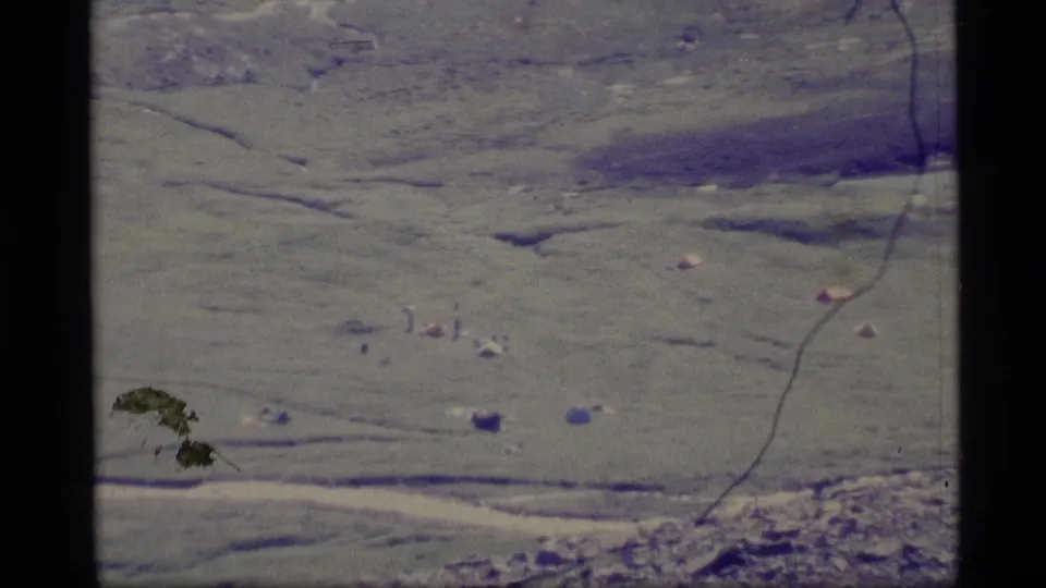1977 Alaska home-movie aerial frame showing green land, small tent-like objects, dark animal-like marks, and a curved track or stream line.
