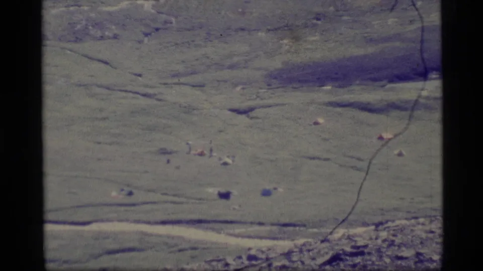 1977 Alaska 8mm aerial still with muted green terrain, small pale tent-like forms, dark marks, and visible grain from the film scan.