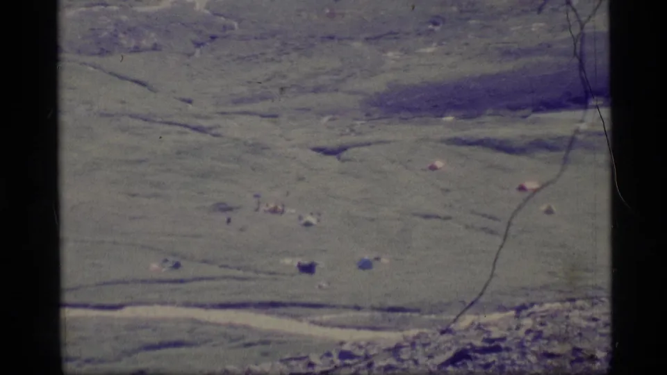 1977 Alaska aerial home-movie frame showing green terrain with small tent-like shapes, dark marks, and a visible vertical film scratch.