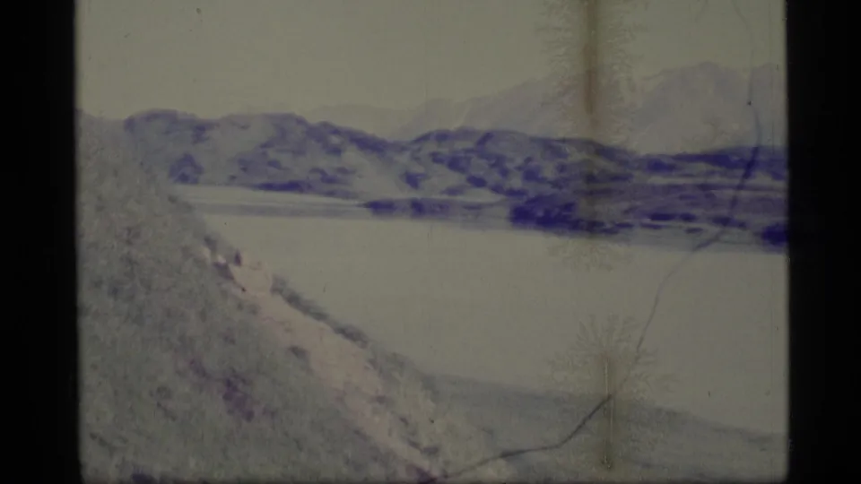 1977 Alaska 8mm frame with a distant mountain ridge, broad pale water, and foreground slope under faded purple-blue film color.