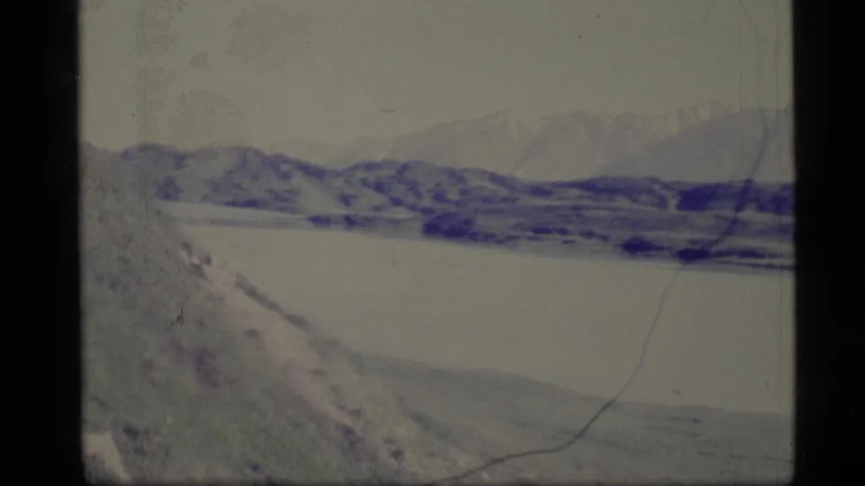 1977 Alaska 8mm frame showing a broad waterway below mountain ridges, with snow or light-toned patches and aged film scratches.