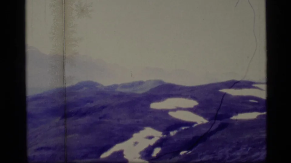 1977 Alaska 8mm frame showing darker mountain slopes, scattered snow patches, hazy distant peaks, and strong purple-blue film shift.