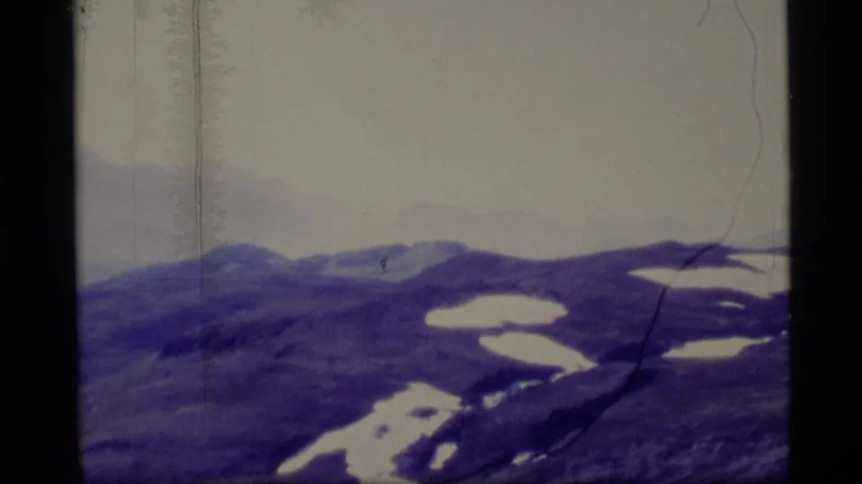 1977 Alaska 8mm frame showing distant snow-patched mountain ridges under a pale sky with visible film gate edges and scratches.