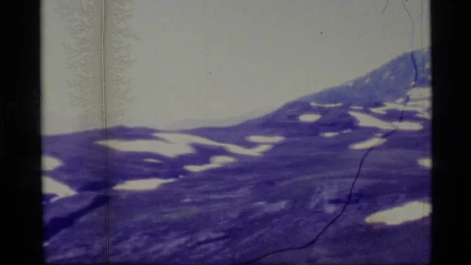 1977 Alaska 8mm frame showing sloped alpine terrain with scattered snow patches and a higher ridge at the right edge.