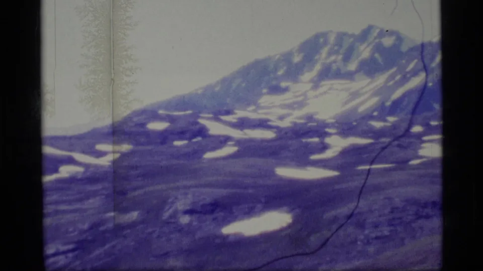 1977 Alaska 8mm frame with a snow-dappled mountain peak, lower alpine slopes, pale sky, and visible scan borders.