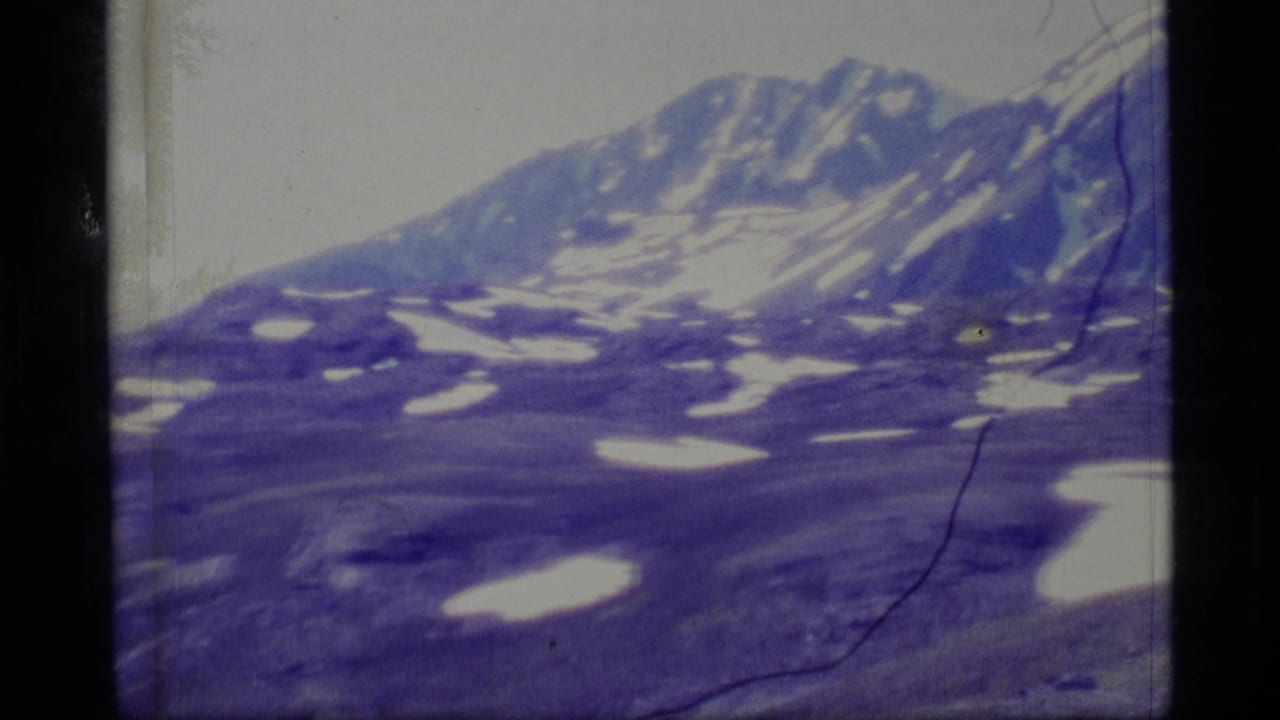 Distant snow-streaked mountain ridge above rocky alpine terrain with visible film scratches.