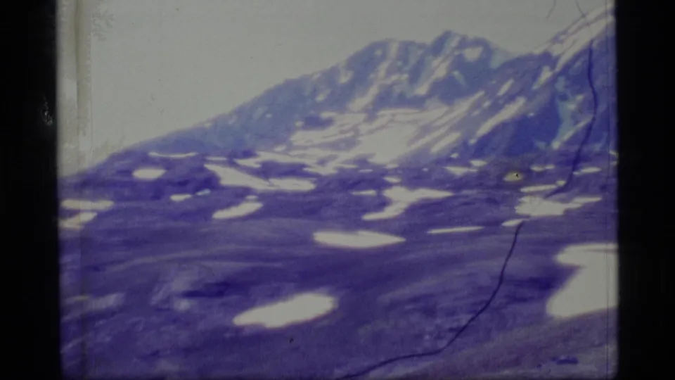1977 8mm Alaska footage showing a distant snow-streaked mountain ridge, rocky slopes, pale sky, and aged blue-purple film color.