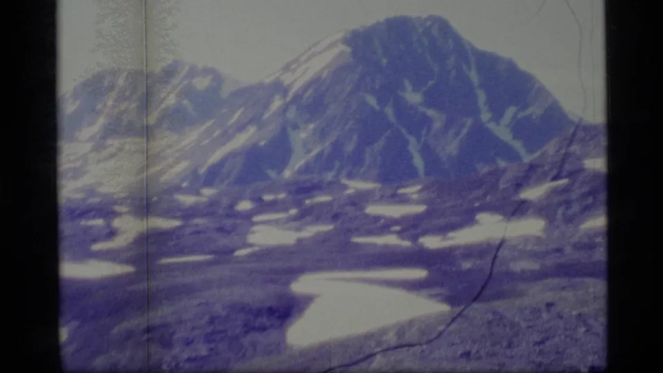 1977 8mm Alaska mountain view with a broad peak, scattered snowfields, rocky foreground texture, and visible film wear.