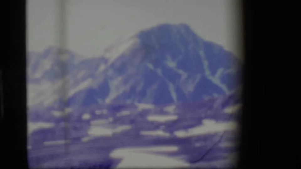 Soft 1977 8mm Alaska frame showing a mountain peak, scattered snow patches, rocky terrain, and heavy blue-purple fading.