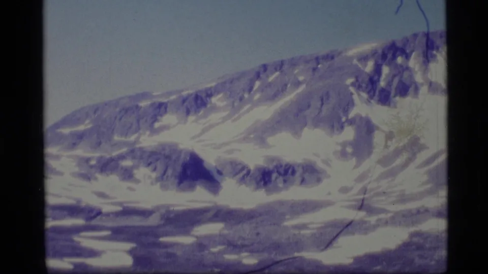 1977 8mm Alaska frame of a long snow-lined mountain slope, exposed rock bands, pale sky, and visible archival film scratches.