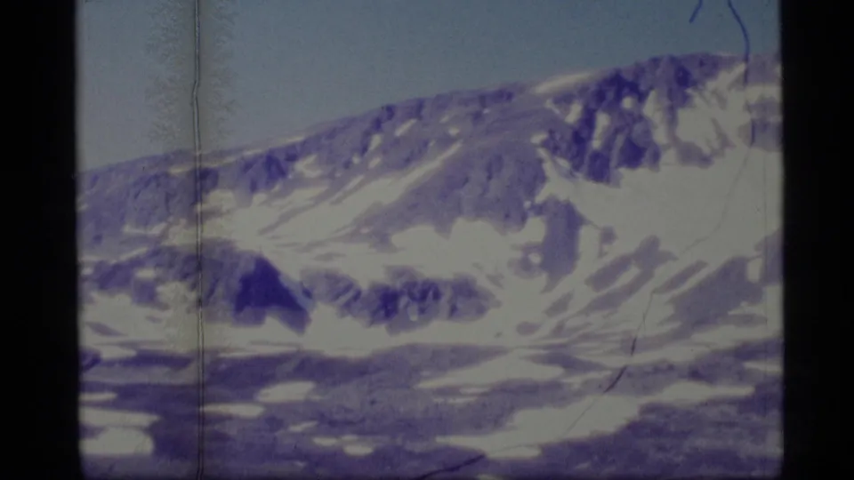 1977 Alaska home-movie frame showing a distant snow-covered mountain face with dark rock bands, purple color cast, and film edge artifacts.