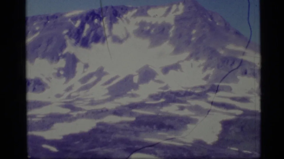1977 Alaska home-movie frame showing a closer snow-streaked mountain face, dark rock shadows, lower valley forms, and aged 8mm film marks.