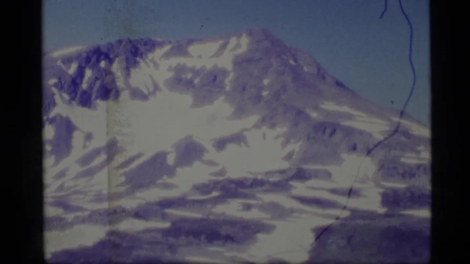 1977 Alaska 8mm frame with a snow-covered mountain peak, sloping ridge, pale blue sky, purple color shift, and visible scan scratches.