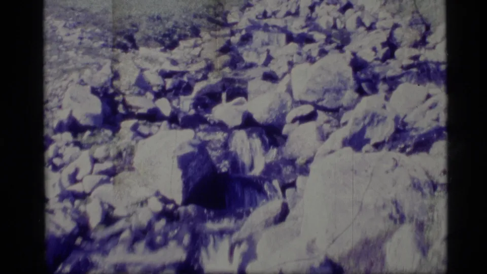 1977 Alaska 8mm view of a rocky stream bed with scattered snow, boulders, and aged blue-white film texture.