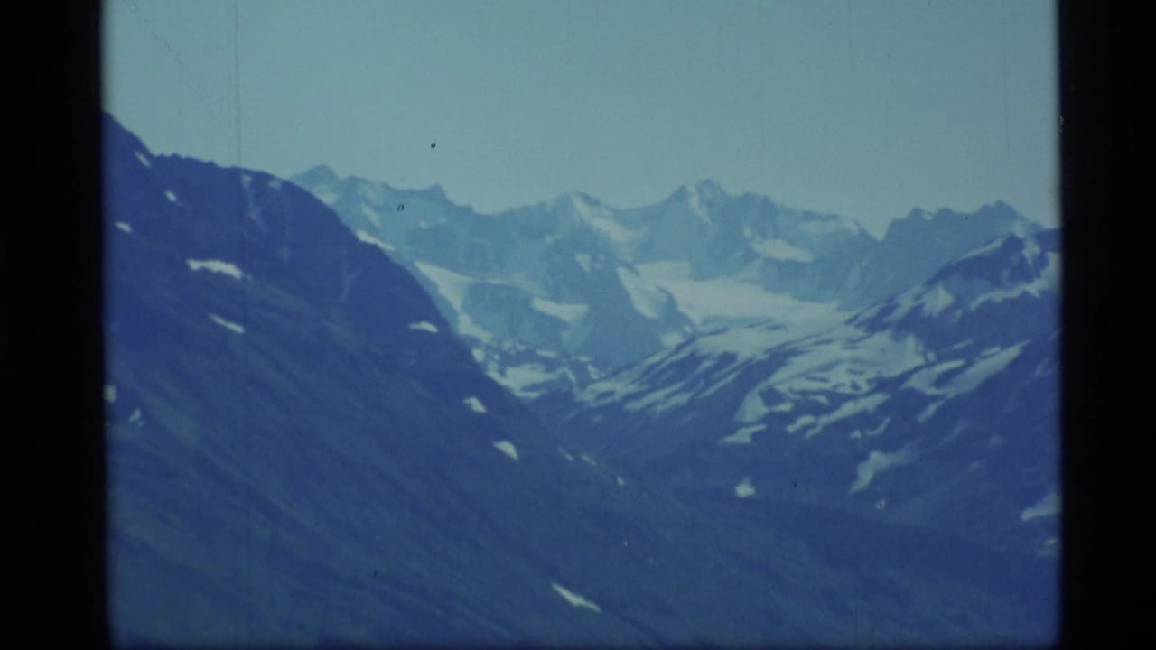 1977 Alaska 8mm frame showing distant snow-capped peaks and dark alpine slopes under a cool blue sky.