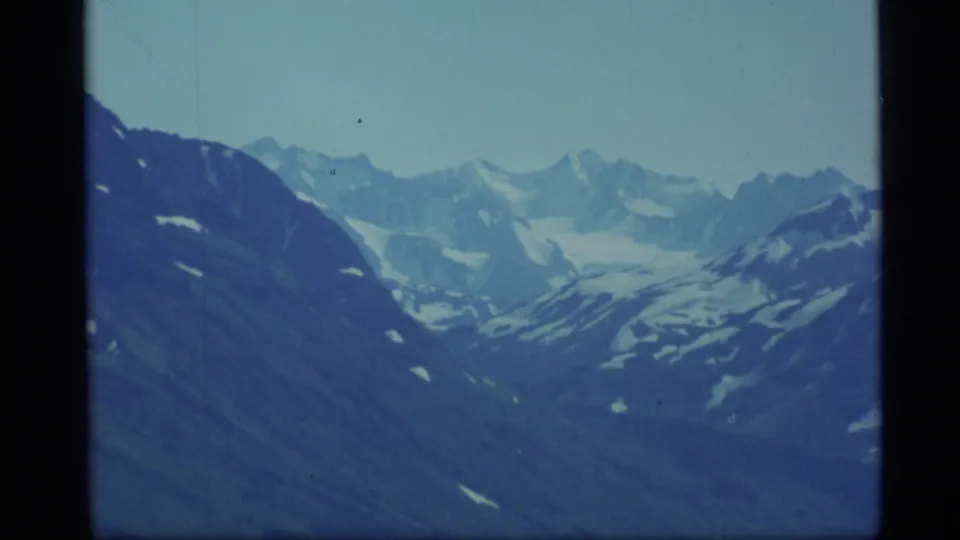 1977 Alaska 8mm frame showing distant snow-capped peaks and dark alpine slopes under a cool blue sky.