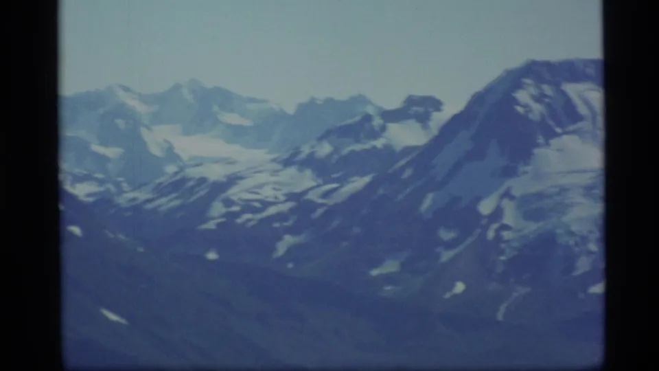 1977 Alaska home-movie frame with snow-covered summits and a broad dark ridge crossing the right side.