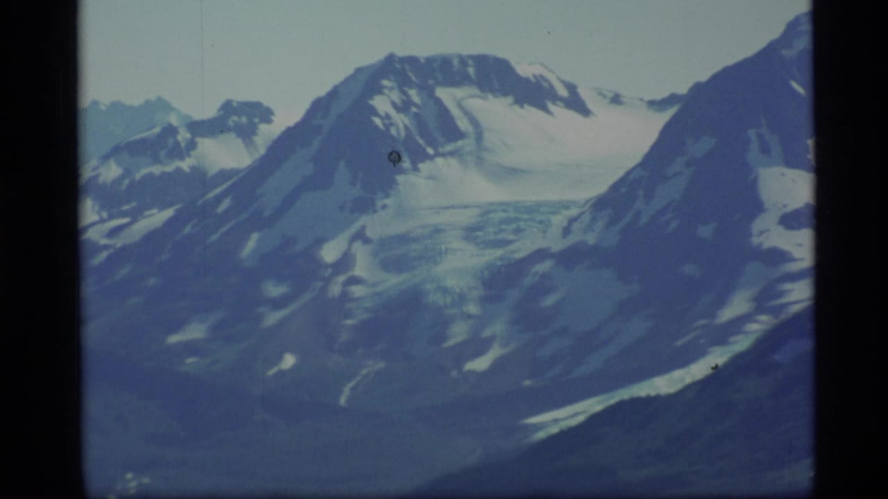 1977 Alaska 8mm frame showing distant snow-covered peaks, glacier-like ice, and dark mountain slopes under a pale sky.