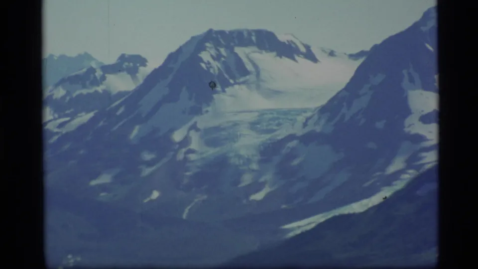 1977 Alaska 8mm frame showing distant snow-covered peaks, glacier-like ice, and dark mountain slopes under a pale sky.