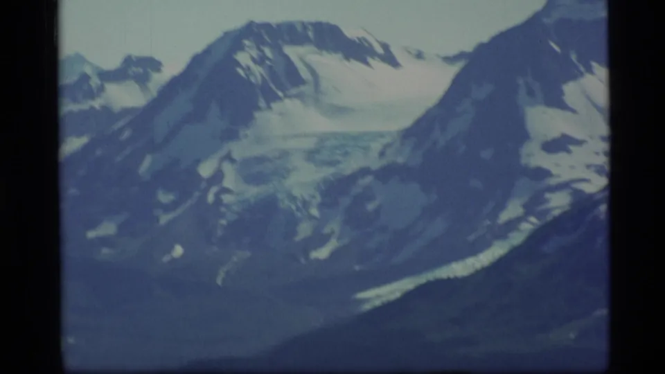 1977 Alaska home-movie frame of icy mountain ridges, broad snowfields, and shadowed slopes during a slow landscape pan.