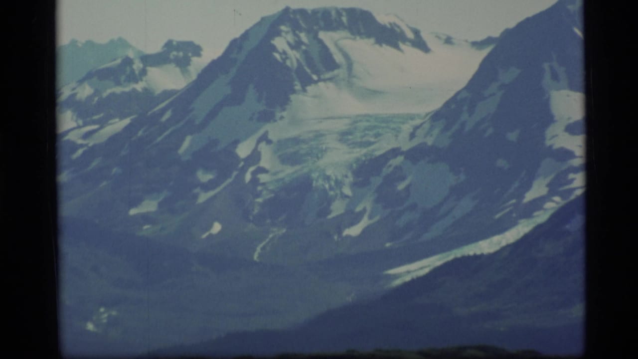 1977 Alaska 8mm frame showing distant snow-covered mountain ridges, glacier-like ice, and a dark green lower slope.