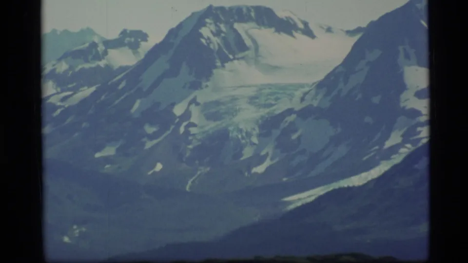 1977 Alaska 8mm frame showing distant snow-covered mountain ridges, glacier-like ice, and a dark green lower slope.
