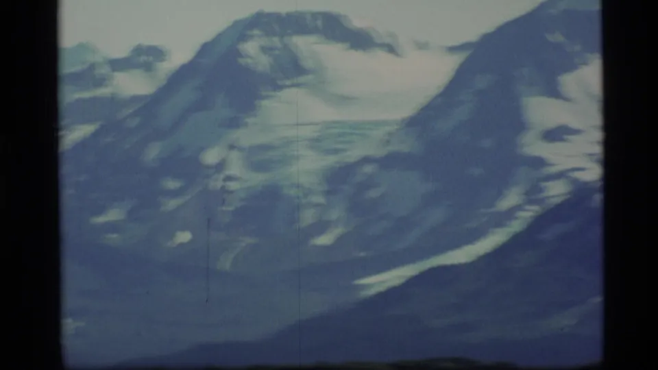 1977 8mm Alaska frame with motion-softened snowy peaks, glacier-like ice, and a dark green foreground.